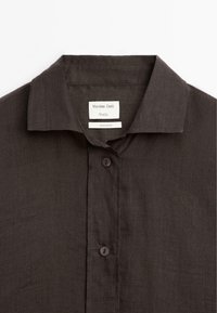 Ciemnobrązowa koszula z guzikiem z klasycznym kołnierzykiem i metką z napisem "Massimo Dutti Studio" oraz "Wyprodukowano w Hiszpanii."