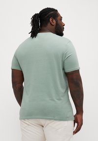 T-shirt a maniche corte, collo rotondo, in tessuto verde chiaro. Texture morbida, vestibilità rilassata e tatuaggi visibili sul braccio.