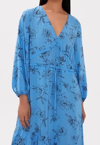 Robe florale bleue avec de longues manches bouffantes, décolleté en V, taille froncée et motifs floraux noirs, fabriquée en tissu léger.