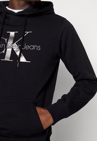 Felpa nera con cappuccio caratterizzata da un grande logo argento "CK" e il testo "Klein Jeans", con cappuccio regolabile e tasca frontale a marsupio. Tessuto morbido con polsini a coste.