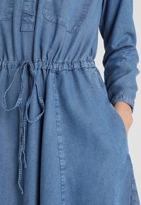 Robe en denim bleu clair, dotée d'une taille resserrée avec un cordon, de manches longues et de deux poches latérales. Texture lisse et coupe décontractée.