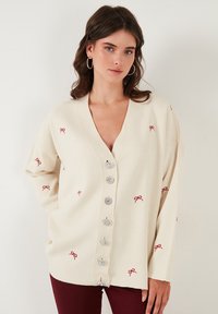 Cardigan crema con scollo a V profondo, decorato con ricami a forma di fiocco rosso e grandi bottoni argentati sulla parte anteriore. Vestibilità rilassata e tessuto texturizzato.