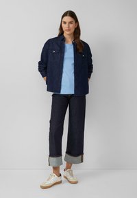 Vrouw die staat met een donkere spijkerjack, lichtblauw shirt, donkere opgerolde spijkerbroek en witte sneakers tegen een effen lichte achtergrond.