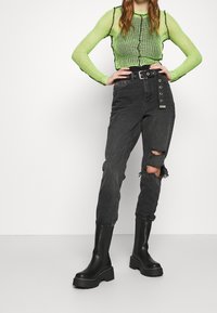 Top de manga larga de malla verde con ribetes negros, combinado con pantalones de mezclilla negros rasgados que presentan detalles de botones de metal y botas negras gruesas.