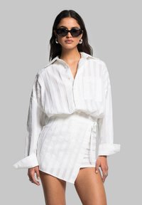 Weiß gestreifte Button-up-Bluse mit lockerer Passform, kombiniert mit einem weißen Wickel-Minirock. Die Accessoires umfassen schwarze Sonnenbrillen und silberne Ohrringe.
