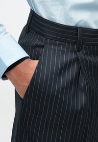 Mão no bolso de calças risca de giz azul-marinho, usadas com uma camisa azul-clara.