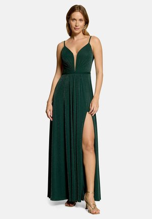Femme portant une robe de soirée sans manches vert foncé avec un profond décolleté en V et une fente haute, associée à des talons hauts dorés à lanières.