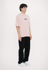 T-shirt oversized rosa com mangas curtas, apresentando um pequeno logotipo preto na frente. Combinado com calças pretas e sapatilhas brancas.