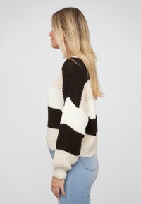 Gestreifter Pullover in Creme- und Dunkelbrauntönen. Grob gestrickte Textur, überschnittene Schultern und gerippte Bündchen. Getragen mit hellblauen Jeans.