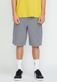 Men's striped shorts in navy and white, featuring a relaxed fit, lightweight fabric, and side pockets. Worn with a yellow t-shirt and black sneakers.
