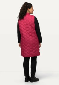Gepolsterte Weste in leuchtendem Pink mit einem hexagonalen Muster, knielangem Design und hohem Kragen, kombiniert mit schwarzen, eng anliegenden Hosen und Kampfstiefeln.