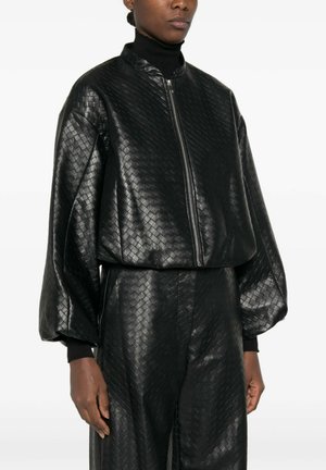 Persoon draagt een zwarte, gestructureerde leren jas met een ritssluiting aan de voorkant en bijpassende hoge taille broek, gecombineerd met een zwarte coltrui eronder.