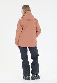 Vesiäskelöity takki pehmeässä korallinvärisessä sävyssä, jossa on huppu, yhdistettynä tummansinisiin housuihin. Takissa on rento malli ja erottuvat vetoketjut.