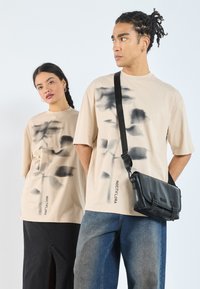 Le t-shirt beige oversize presenta motivi astratti neri e il testo "NOCTIFLORA" sul lato. Il modello maschile porta una borsa a tracolla nera.