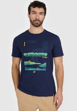 Mężczyzna z brodą, ubrany w granatowy t-shirt z zielonym wzorem gór i napisem „OUTDOORS est. 1995”, beżowe spodnie, stoi z ręką w kieszeni.