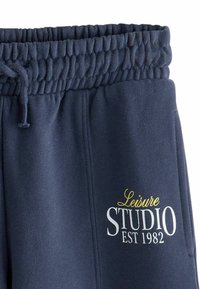 Un pantalon de jogging bleu marine avec une taille élastique, un cordon de serrage, et un texte graphique blanc et or indiquant "Leisure STUDIO EST 1982."