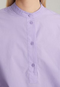 Camicia button-up lavanda con colletto a banda, caratterizzata da due bottoni e un tessuto leggermente texturizzato. Design semplice senza motivi.