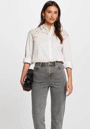 White button-up shirt with lace detailing on shoulders, paired with high-waisted gray jeans. Model is holding a small black bag.