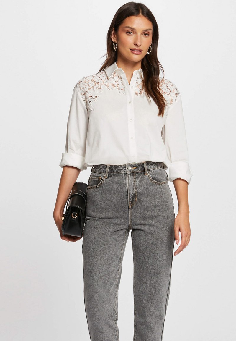 Camicia bianca con bottoni e dettagli in pizzo sulle spalle, abbinata a jeans grigi a vita alta. La modella tiene in mano una piccola borsa nera.