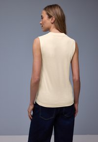 Femme aux cheveux longs châtain clair, portant un haut crème sans manches et un jean bleu foncé, debout de profil sur un fond gris uni.