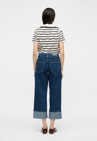 Γυναίκα που φοράει κοντομάνικη ριγέ πλεκτή μπλούζα, ψηλόμεσα φαρδιά cropped τζιν με γυρισμένα ρεβέρ και καφέ φλατ παπούτσια, στέκεται γυρισμένη με την πλάτη.