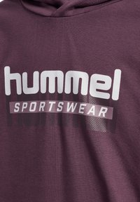 Donkerpaarse stof met "hummel" in grote witte letters en "SPORTSWEAR" in kleinere witte letters eronder.