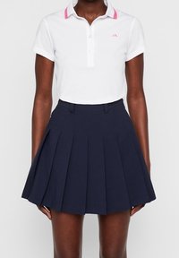 Kvinna klädd i en vit kortärmad polo med rosa krage och en högmidjad marinblå plisserad kjol.