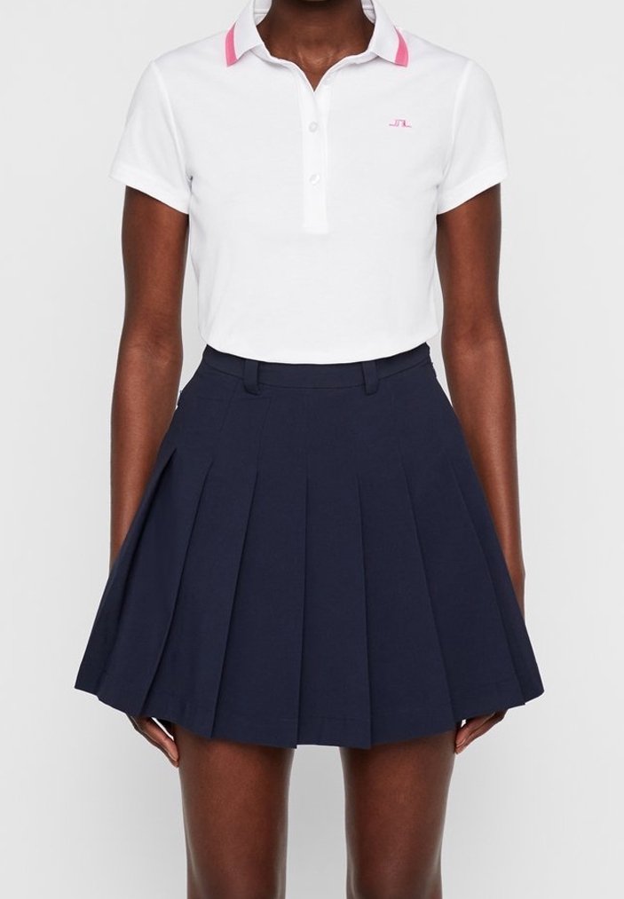 Kvinna klädd i en vit kortärmad polo med rosa krage och en högmidjad marinblå plisserad kjol.