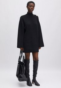 Schwarzes, gestricktes, übergroßes Sweaterkleid mit hohem Kragen, großen Ärmeln und goldenen Knopfdetails an der Schulter, kombiniert mit einer glänzenden schwarzen Tote-Tasche.