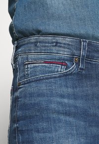Detailní záběr modrých džínsů ukazující zadní kapsu se červeným a bílým prošíváním a detaily nýtů.