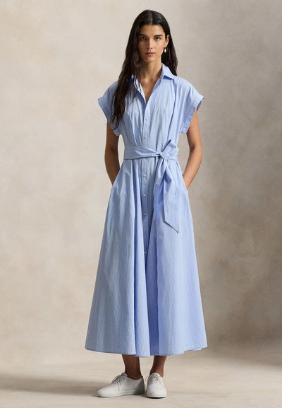 Φόρεμα maxi πουκάμισο με γαλάζιες ρίγες, κοντά μανίκια καπέλο, κλείσιμο με κουμπιά, ζώνη στη μέση και φαρδιά φούστα. Συνδυασμένο με λευκά παπούτσια.