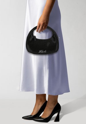 Bolso de mano negro con cuentas, con un asa curva y cierre de cremallera, que presenta un detalle de logo en plata, sostenido por una persona que lleva un vestido de satén blanco y tacones.