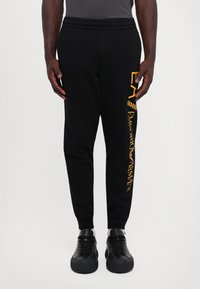 Čierne bavlnené nohavice typu joggers s elastickým pásom, so žltým potlačeným logom pozdĺž ľavej nohy. Úzky strih s rebrovanými manžetami na členkoch.