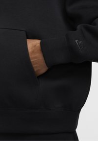 Černá mikina s přední kapsou a žebrovanými manžetami, s nenápadným černým logem Nike na rukávu. Vyrobena z měkké, texturované látky.