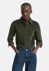 Woman with braided hair wearing a dark green button-up shirt with rolled sleeves and high-waisted blue jeans, hands in pockets.