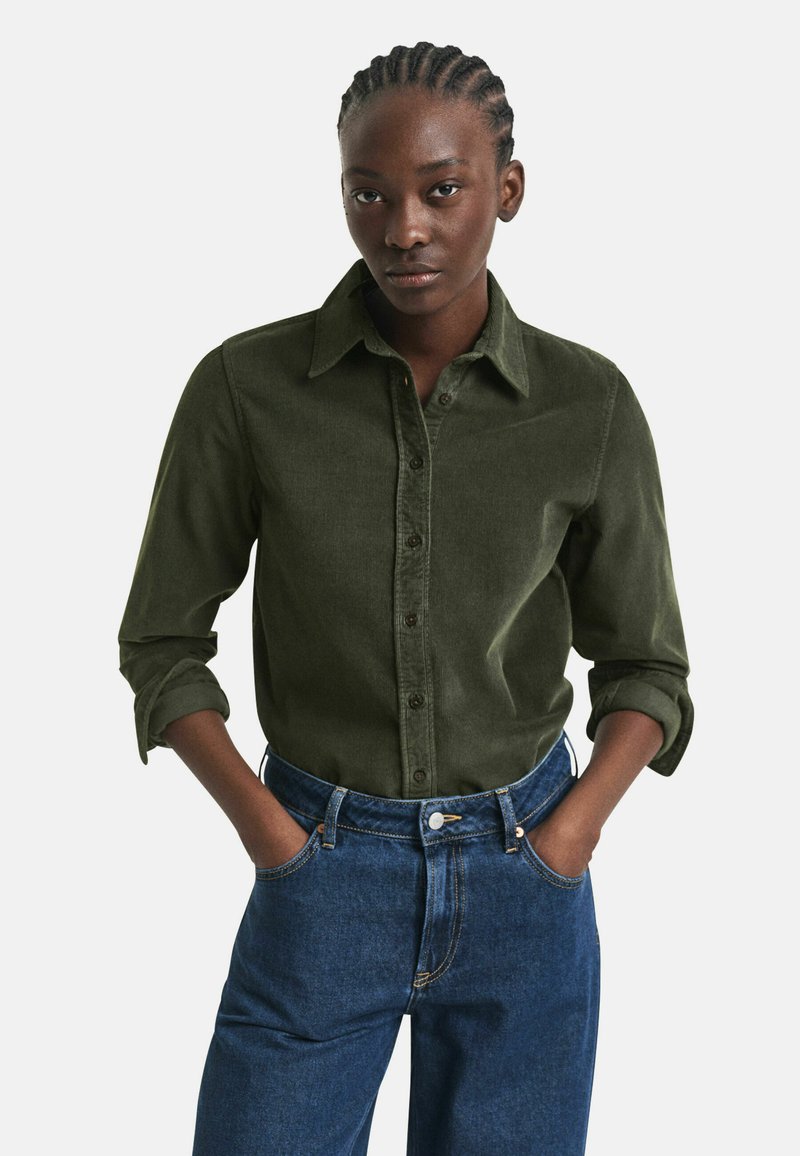 Woman with braided hair wearing a dark green button-up shirt with rolled sleeves and high-waisted blue jeans, hands in pockets.