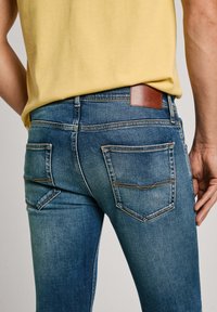 Jeans in denim blu con una vestibilità slim, dotati di due tasche posteriori con cuciture beige e un'etichetta del marchio in pelle marrone sulla cintura.