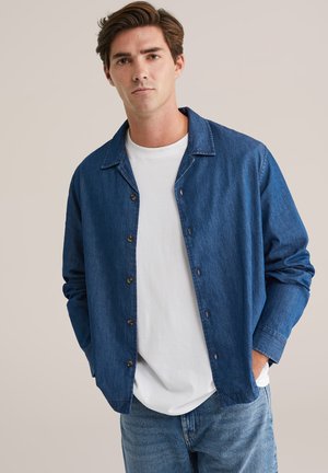 Jeans jakna v temno modri barvi z mehkimi teksturami, z ovratnikom, sprednjimi gumbi in dolgimi rokavi, oblečena čez belo majico.