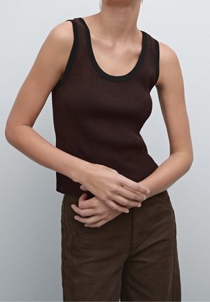 Personne portant un haut sans manches côtelé marron foncé avec bordure noire et un pantalon marron, les mains doucement jointes devant.