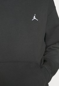 Sweat-shirt noir avec logo Jumpman blanc brodé sur la poitrine, poche avant visible, et une main dans la poche.