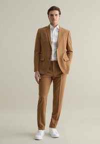 Hellbrauner Anzug mit zwei Knöpfen, kombiniert mit einem weißen Hemd. Slim Fit, glatte Textur, getragen mit weißen Sneakers für einen lässigen Akzent.