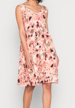 Robe sans manches longueur genou avec motif floral rose, beige et noir, portée par une femme debout les mains reposant le long du corps.
