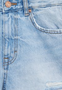 Jean en denim bleu clair en gros plan montrant la poche avant, les rivets en cuivre, la boucle de ceinture et une petite déchirure usée près du coin inférieur droit.