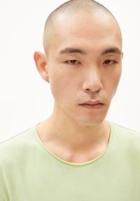 Hellgrünes Baumwoll-T-Shirt mit Rundhalsausschnitt; das Model hat einen rasierten Kopf und einen kleinen Hängeohrring; neutraler Hintergrund.