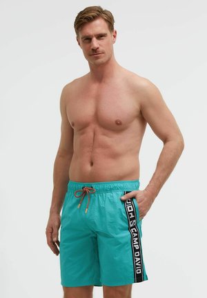 Oberkörperfreier Mann mit hellbraunen Haaren, der türkisfarbene Badeshorts mit einem schwarzen und weißen "Camp David"-Seitenstreifen trägt, mit der Hand in der Tasche.