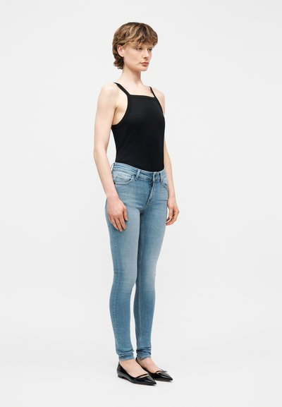 Νεαρή γυναίκα με κοντά μαλλιά που φοράει μαύρο τοπ με τετράγωνη λαιμόκοψη, ανοιχτό μπλε στενά τζιν και μαύρες μυτερές μπαλαρίνες, στέκεται μπροστά σε λευκό φόντο.