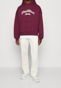 Person som bär en vinröd hoodie med texten "WnstBhv Studio", vita byxor och vita sneakers, stående mot en ljus, enfärgad bakgrund.