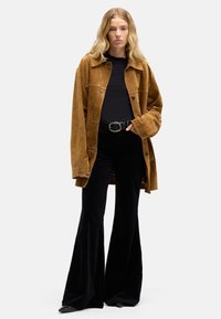 Giacca oversize in camoscio marrone con colletto, camicia aderente nera e pantaloni neri svasati. Caratterizzata da una cintura nera con fibbia rotonda in metallo.