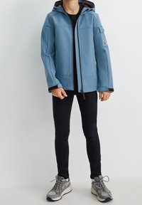 Niebieska kurtka typu softshell z kapturem, zapinana na zamek, z czarnymi wykończeniami i dwiema przednimi kieszeniami. Noszona na czarną bluzkę, ciemne dżinsy i sneakersy.