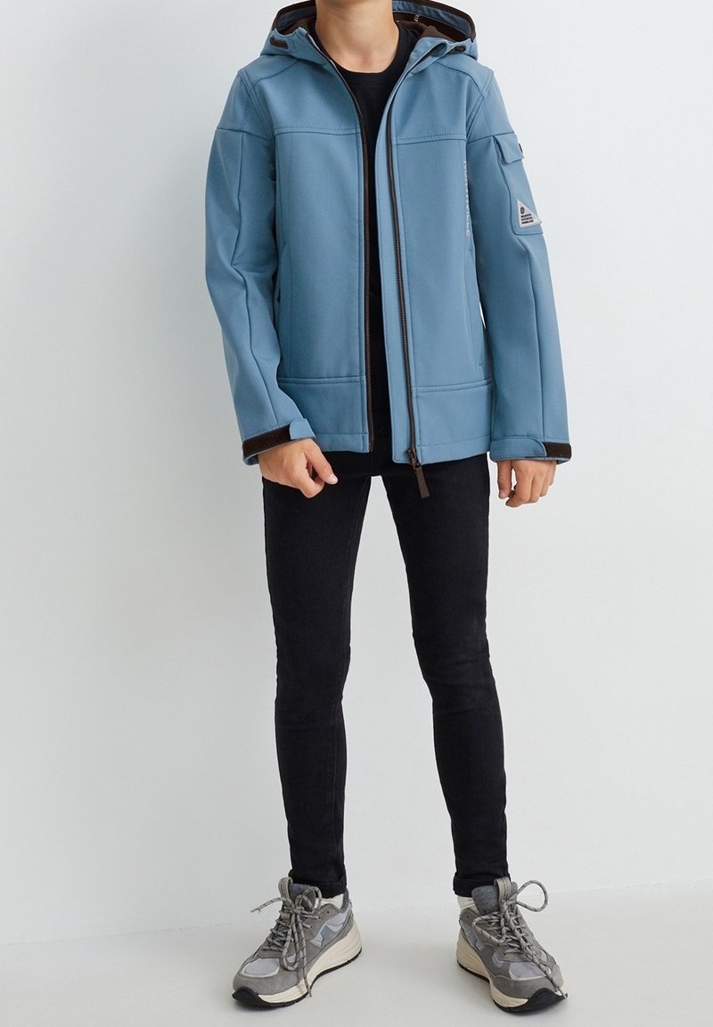 Niebieska kurtka typu softshell z kapturem, zapinana na zamek, z czarnymi wykończeniami i dwiema przednimi kieszeniami. Noszona na czarną bluzkę, ciemne dżinsy i sneakersy.